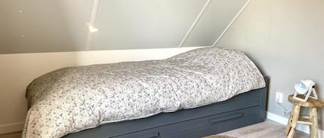 Un lit simple recouvert d’une couvre-lit à motifs floraux, un tiroir de couleur sombre en dessous, un tapis tissé sur le sol et un petit tabouret en bois avec une lampe blanche à côté du lit.