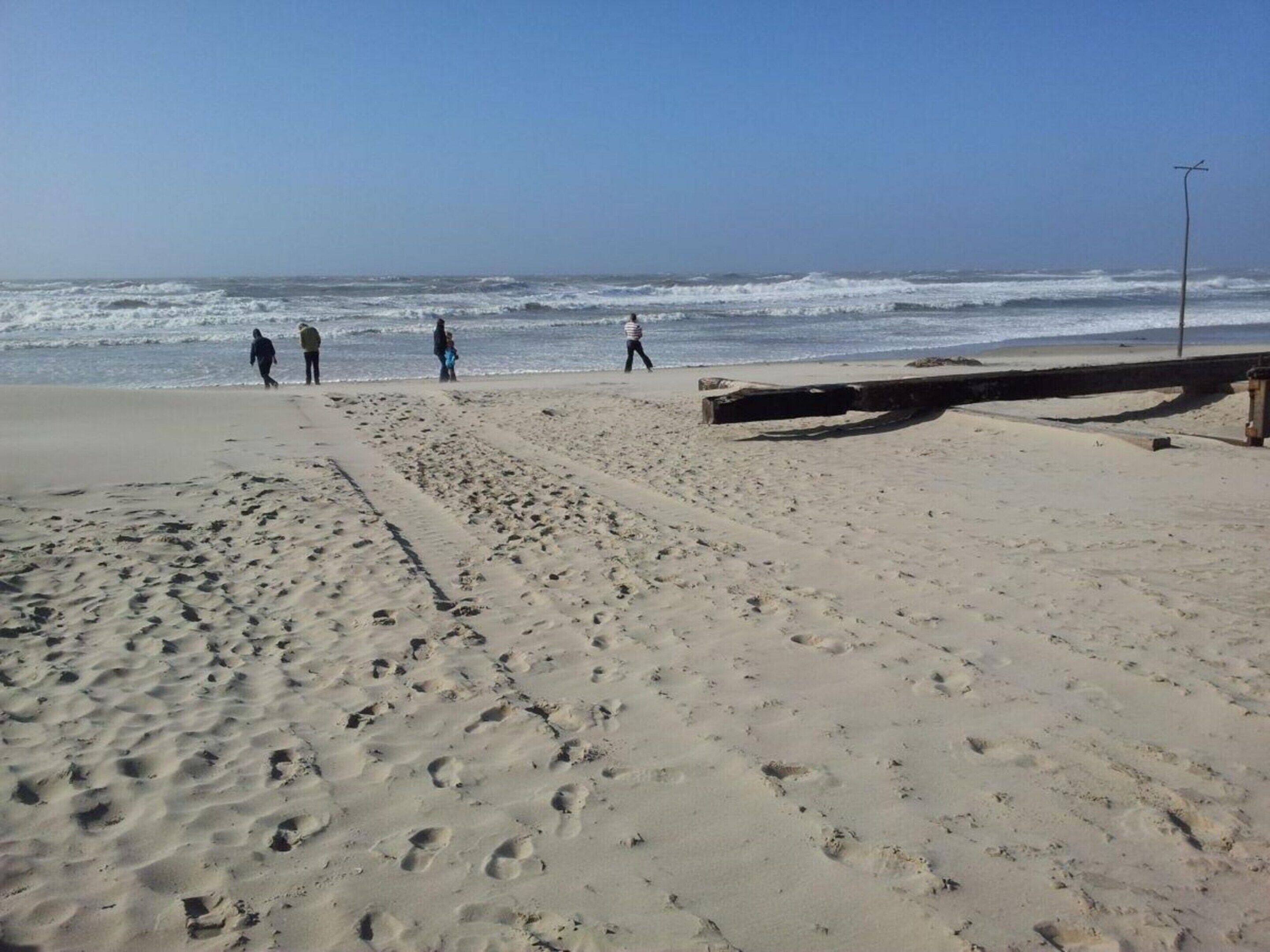 A sandy beach with footprints leading to the water, a wooden structure, and a clear sky.