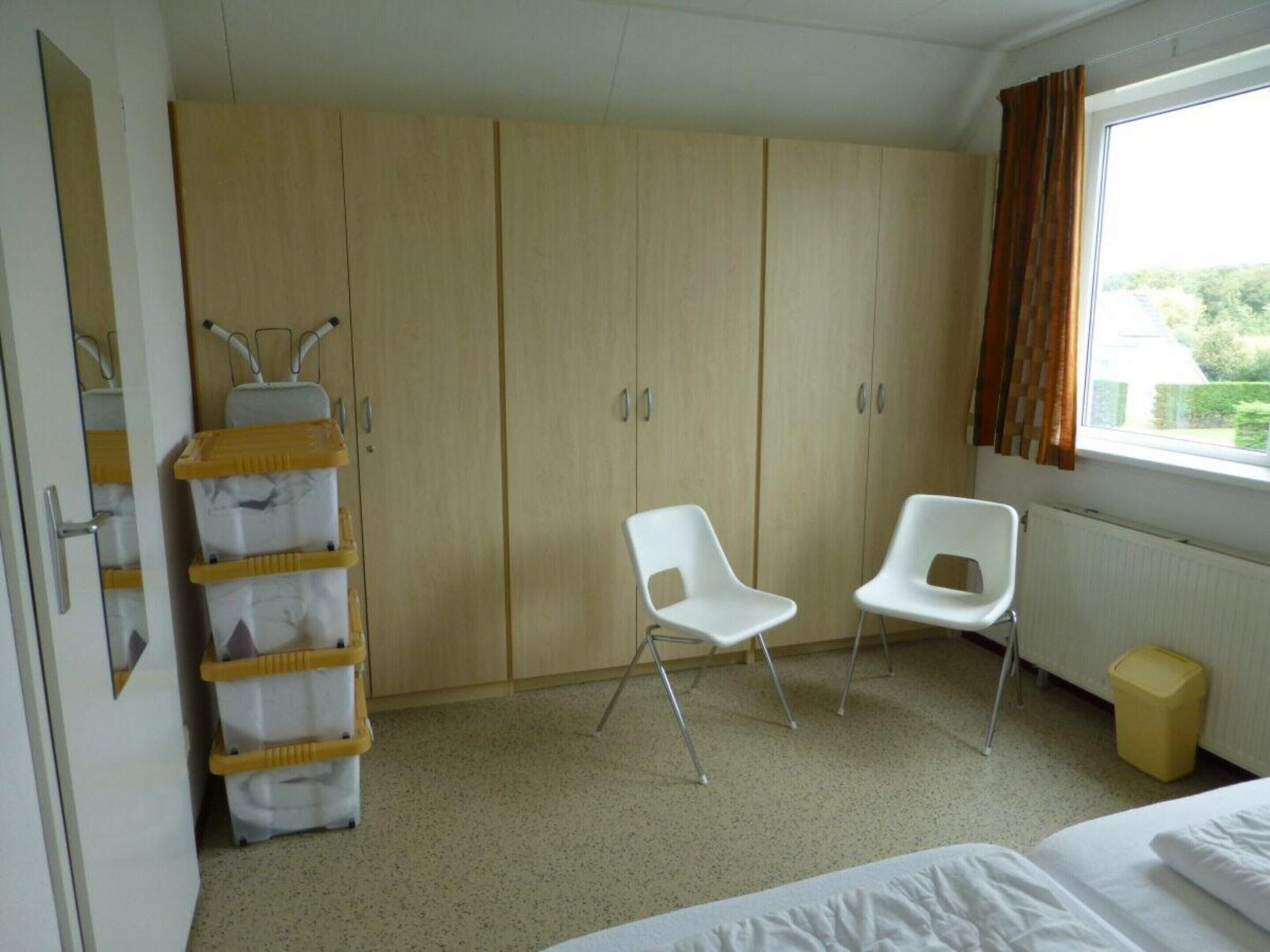 A room with a bed, two white chairs, a wooden wardrobe, a radiator, and a window with curtains.