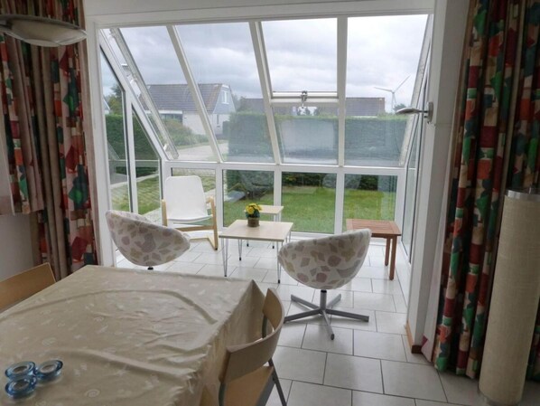 A room with a large glass door leading to a garden, a table with a tablecloth, two patterned chairs, and a small table with a vase of flowers.