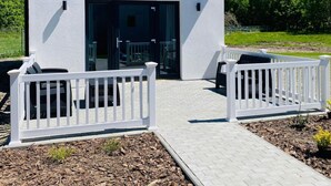 Una casa moderna con fachada blanca, techo negro y un amplio ventanal. Se ven dos cercas blancas con sillas al frente.