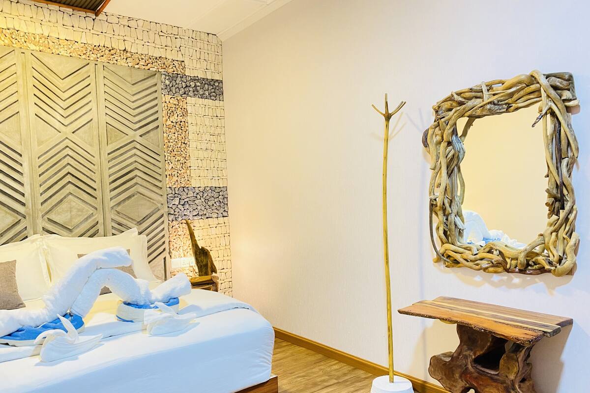Un lit bien fait, avec du linge de lit blanc et un coussin décoratif en forme de cygne, un miroir fixé au mur et une table de chevet en bois rustique.