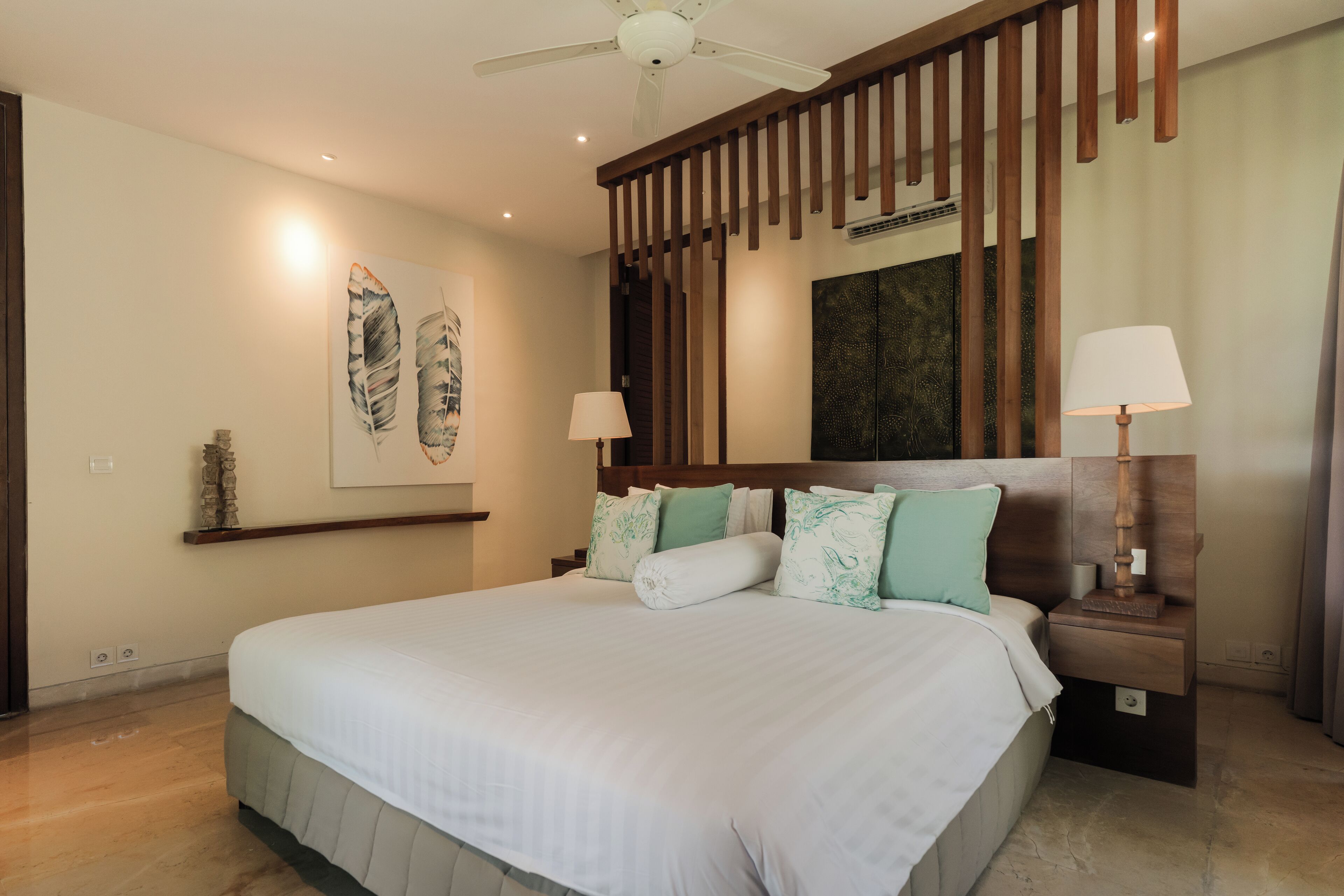 A spacious bedroom with a large bed, wooden headboard, two bedside tables with lamps, a ceiling fan, and artwork on the walls.