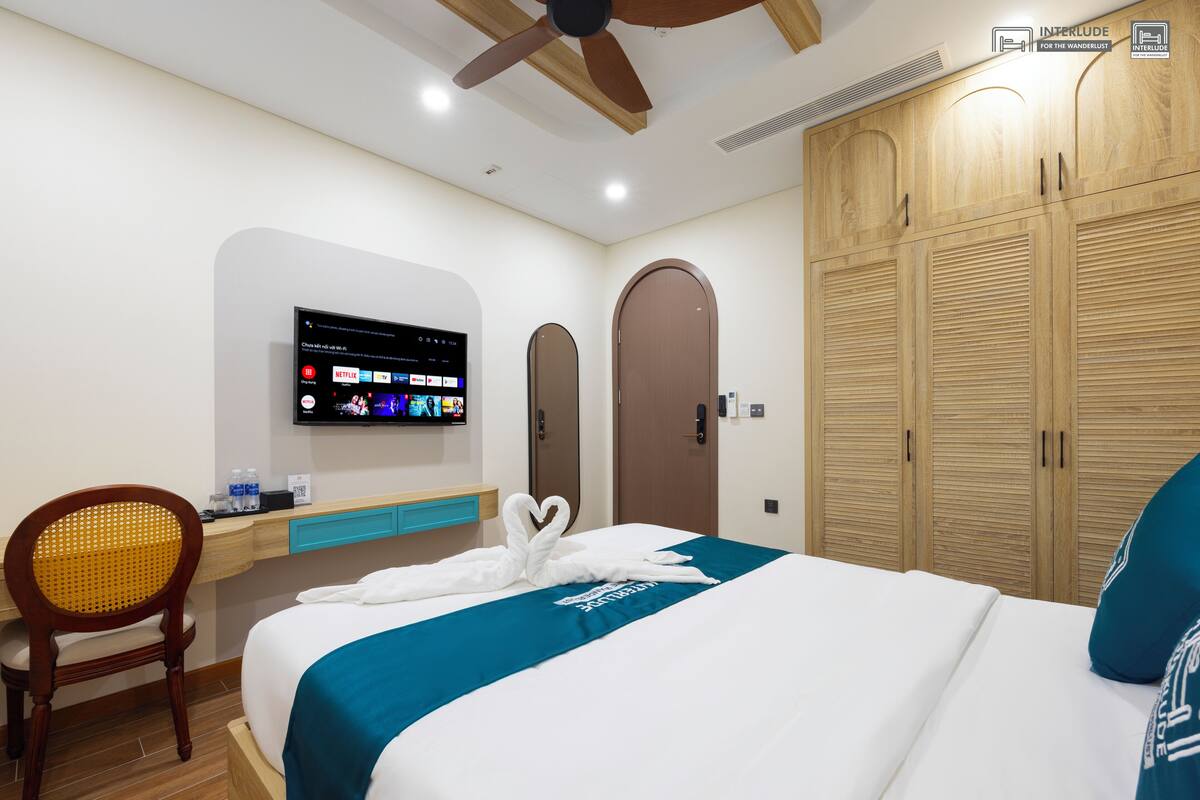 A hotel room with a bed, a TV mounted on the wall, a wooden wardrobe, and a ceiling fan.