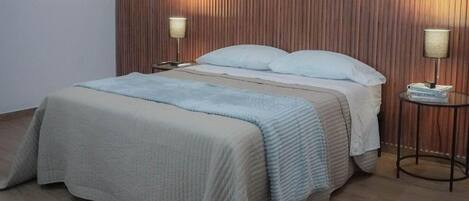 Una cama bien hecha con edredón y almohadas de color claro, flanqueada por dos mesitas de noche con lámparas. La habitación tiene paneles de madera en las paredes y piso de madera.
