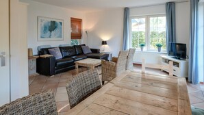 Un salon avec un canapé en cuir noir, une table à manger en bois et des chaises en osier, un téléviseur posé sur un meuble blanc, et une fenêtre avec des rideaux.