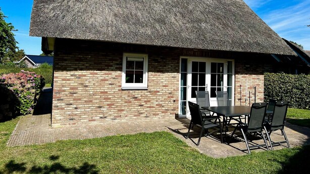 Un charmant cottage à toit de chaume, avec une façade en briques, un petit patio doté d’une table et de chaises, et un jardin bien entretenu.