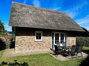 Un charmant cottage à toit de chaume, avec une façade en briques, un petit patio doté d’une table et de chaises, et un jardin bien entretenu.