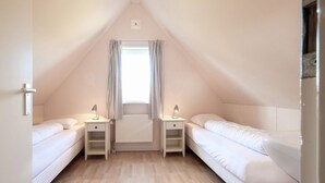Une petite chambre mansardée triangulaire, avec deux lits simples, une fenêtre avec des rideaux et un radiateur.