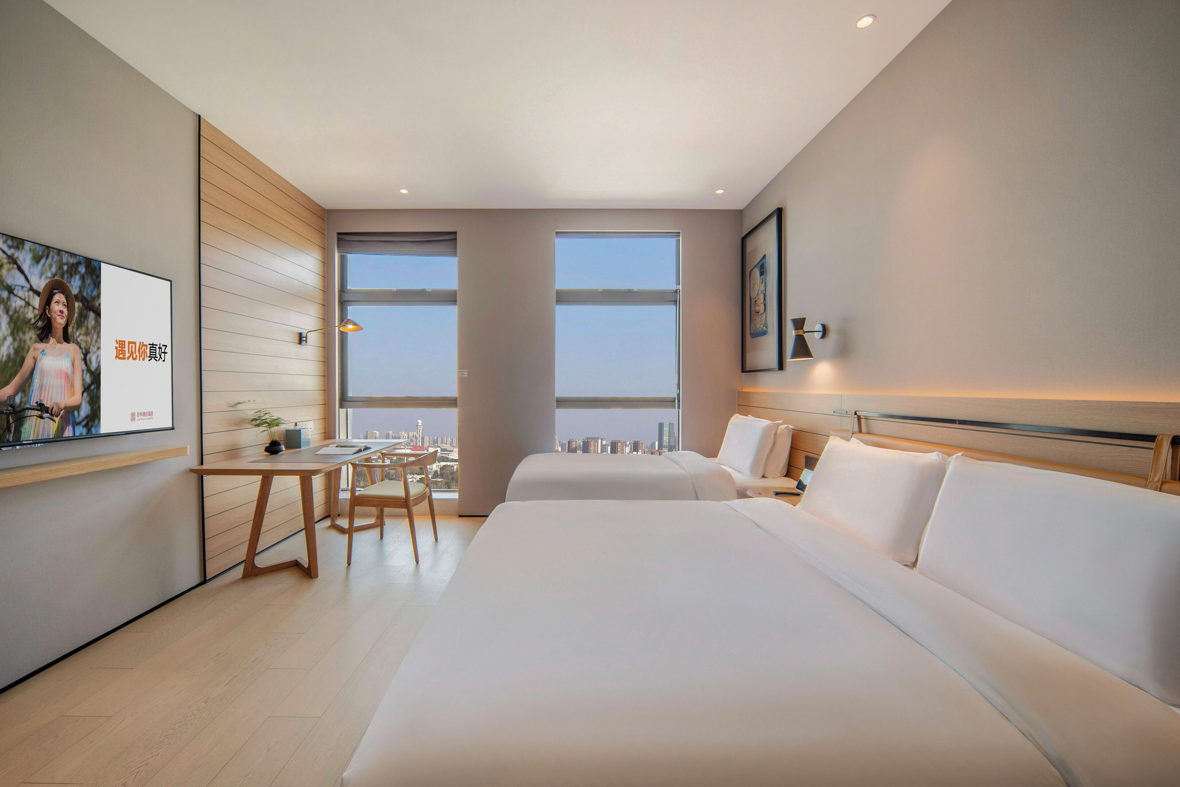 A modern hotel room with a large bed, a desk, and a chair. There are two windows providing a view of the cityscape.