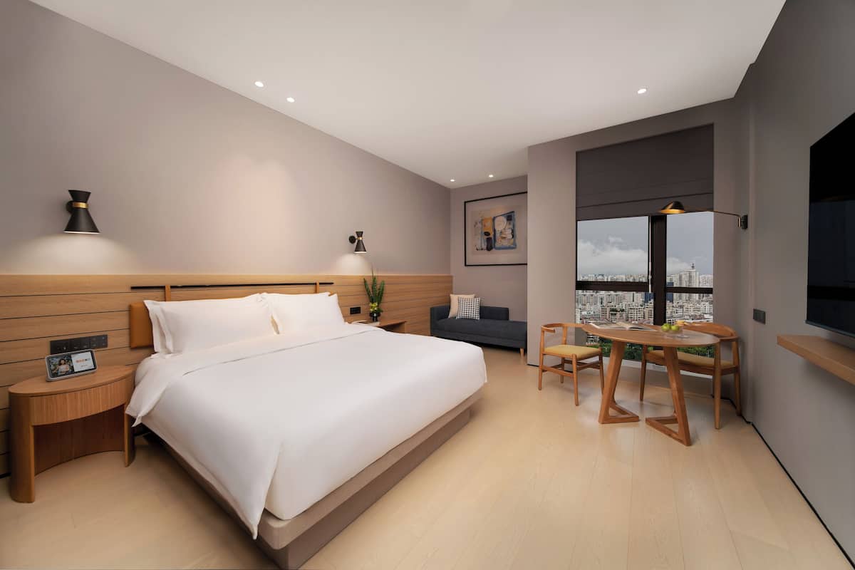 Een moderne hotelkamer met een groot bed, nachtkastjes, een bureau met een stoel, een bank en uitzicht op de stad door een groot raam.