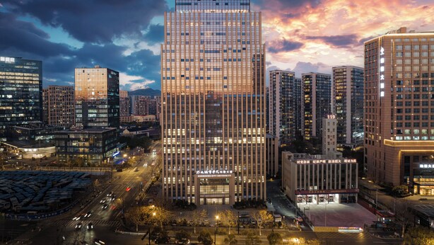 Een modern hotelgebouw met een gegrid façade, omringd door andere hoogbouw en een drukke straat in de schemering.