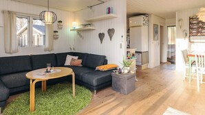 Ein modernes Wohnzimmer mit einem dunkelgrauen Ecksofa, einem runden Holztisch und einem grünen Hochflor-Teppich. Es gibt eine Küchenzeile mit einem weißen Kühlschrank sowie einen Esstisch mit Stühlen.
