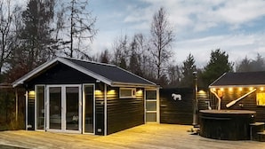 Eine Holzterrasse mit überdachtem Außenbereich, einem Whirlpool und einem kleinen Gebäude mit Glasüren.