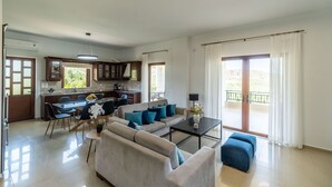 A modern living room with a sofa, coffee table, and dining area. There is a balcony with a view of the landscape.