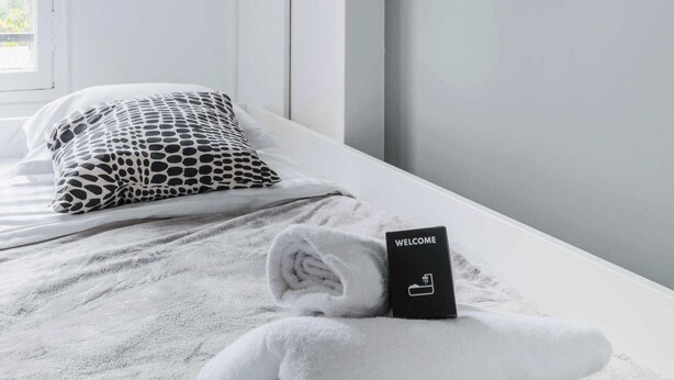 Una cama bien hecha con una toalla doblada y un cartel de bienvenida, una lámpara de pared y una ventana con luz natural.
