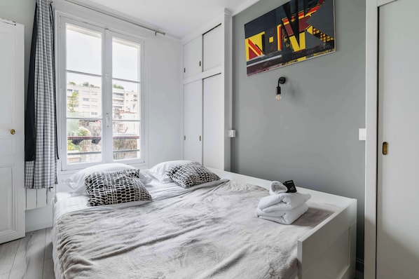 A bedroom with a bed, pillows, a blanket, a towel, a window, a door, and a wall with a painting. - Studio Cosy proche tour Eiffel - Paris 7 - 2P (Paris)