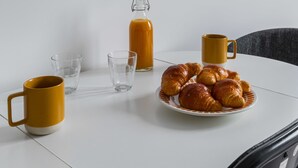 Una mesa blanca con un plato de croissants, una botella de vidrio y dos tazas amarillas.