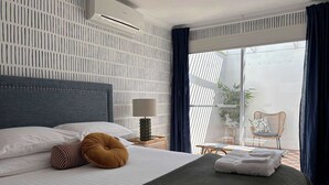 Ein modernes Hotelzimmer mit einem großen Bett, einem Nachttisch, einer Lampe und Blick auf einen Balkon mit Stuhl und Pflanzen.