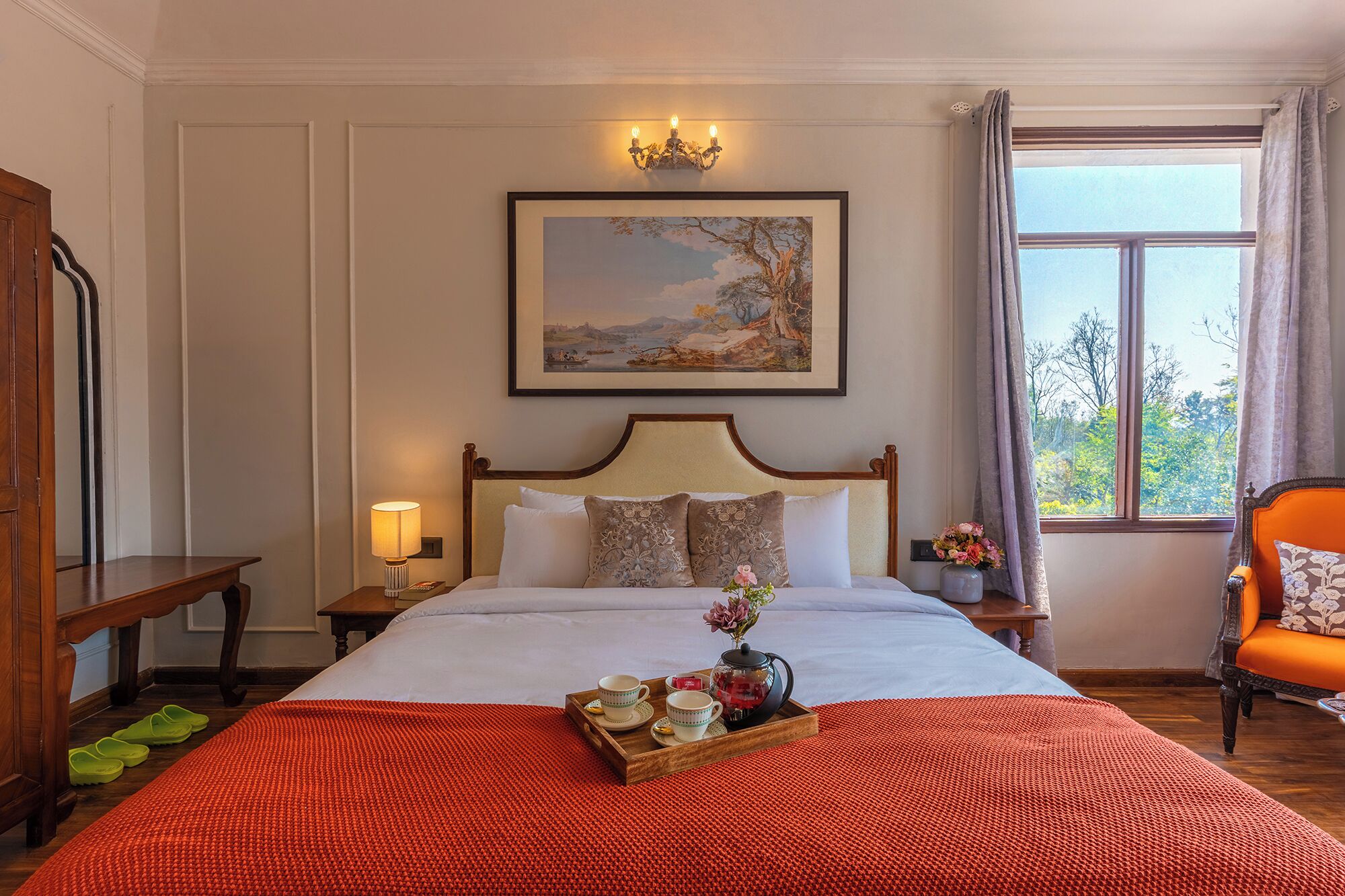 Habitación de hotel impecable con una cama grande, una mesita de noche con lámpara, un cuadro enmarcado sobre la cama, una ventana con cortinas y una silla con cojín floral.