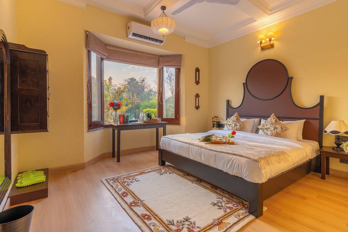 A bedroom with a large bed, a bedside table, a nightstand, a window with curtains, an air conditioning unit, and a view of the outdoors.