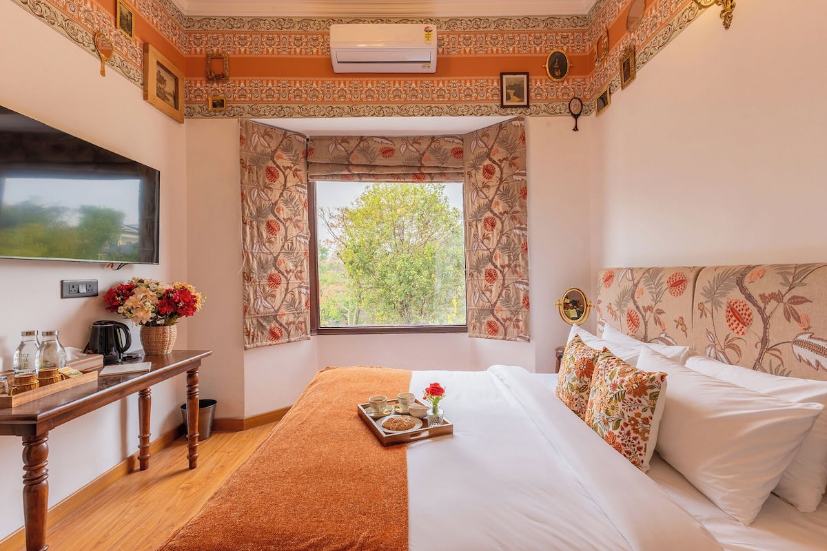 A hotel room with a bed, a wooden table with a tray of breakfast items, a TV, and a window with floral curtains.