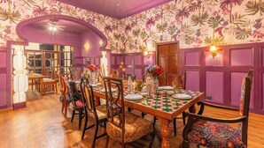 Une salle à manger avec une table en bois dressée pour quatre personnes, entourée de chaises rembourrées. Les murs sont décorés d’un papier peint à motifs floraux.