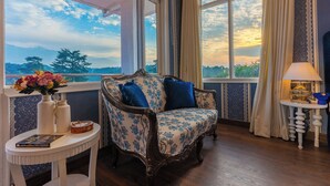 Une pièce offrant une vue sur les montagnes et sur le coucher de soleil, visible à travers de grandes fenêtres, avec un canapé à motifs floraux, une petite table sur laquelle sont posés un vase et des livres, et une lampe sur une table d’appoint.