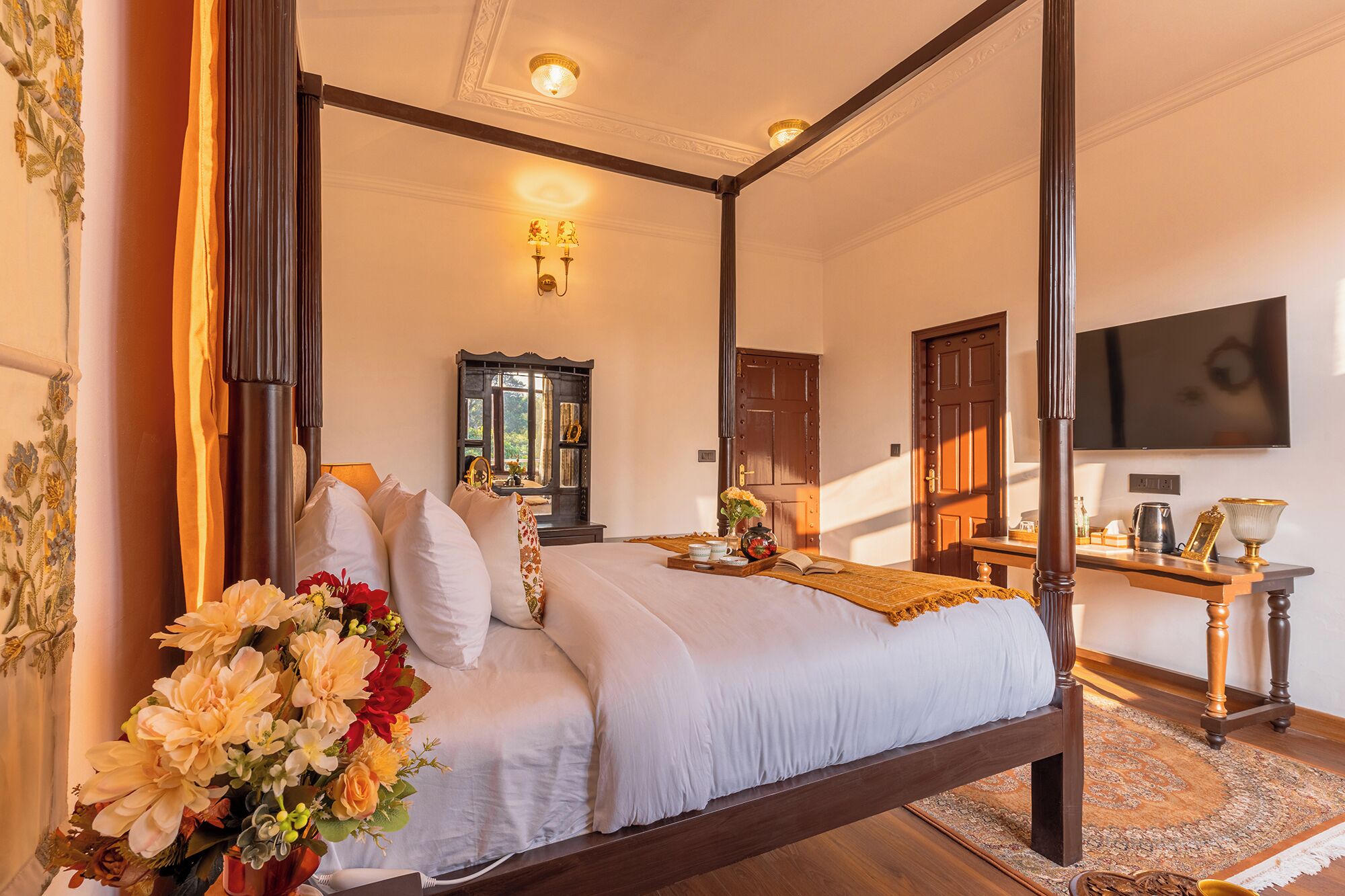 A four-poster bed with white linens, a wooden floor, a large mirror, and a television on the wall.