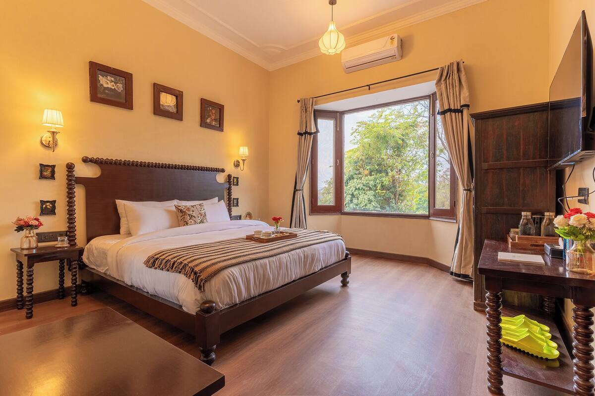 A bedroom with a large bed, wooden headboard, bedside tables, a nightstand, a desk, a chair, a window with curtains, and an air conditioning unit.