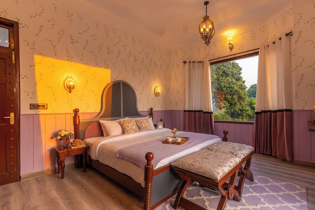 A bedroom with a large bed, a wooden bench, a nightstand, and a window with curtains.