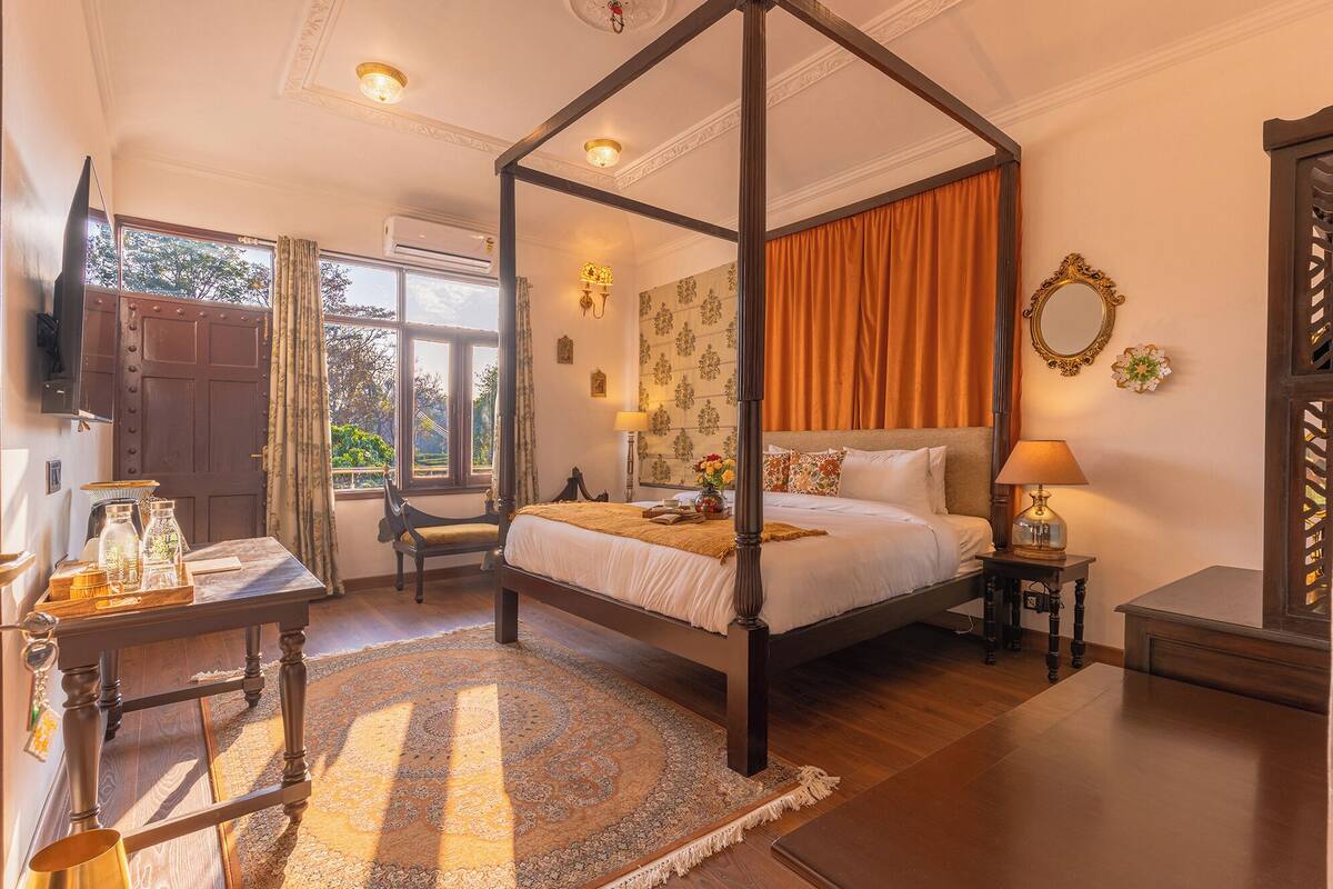 A four-poster canopy bed with a wooden headboard, a bedside table with a lamp, and a view of the outdoors through a window with curtains.