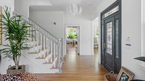 Un couloir avec un escalier, un lustre, une porte sombre à panneaux de verre et, au fond, un coin repas.