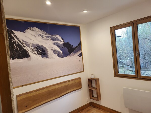 Een kamer met een groot berglandschap op het doek, een houten bankje, een klein houten plankje en een raam met uitzicht op bomen.