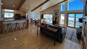 A spacious living area with a wooden ceiling, a kitchen with wooden cabinets, a dining area, and large windows offering a view of the outdoors.