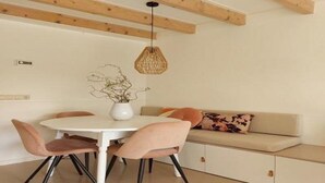 Een eetgedeelte met een ronde tafel, vier stoelen, een vaas met takken, een geweven hanglamp en een bank met kussens.