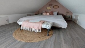 Een slaapkamer met een schuine kap, een bed met een wit en roze dekbedovertrek, een geweven vloerkleed en een hanglamp.
