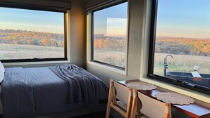 Une chambre à coucher comprenant un lit, un bureau avec une chaise et de grandes fenêtres donnant sur un paysage boisé.