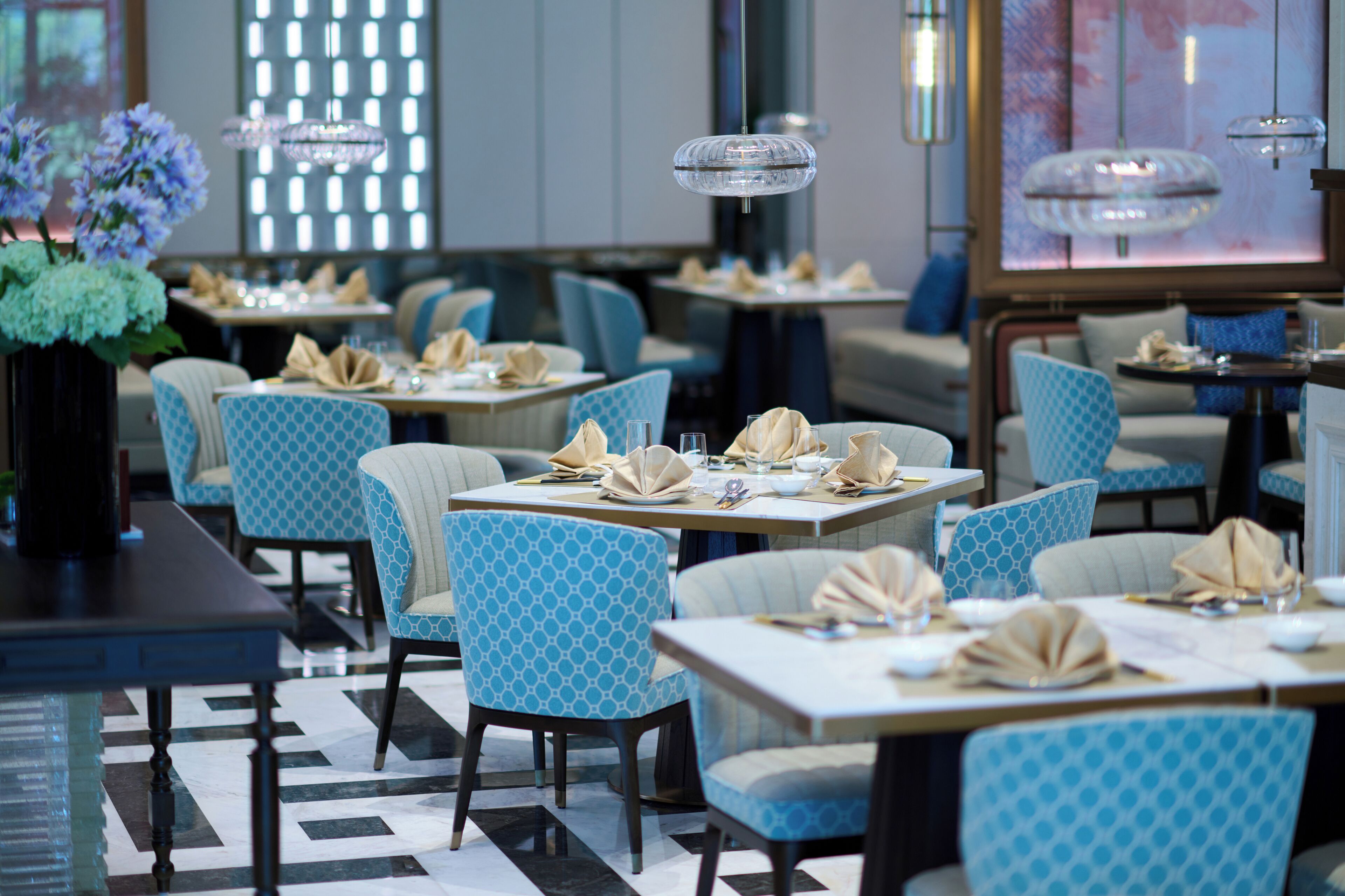 A restaurant with blue patterned chairs, tables set with white tablecloths, and clear glass pendant lights.