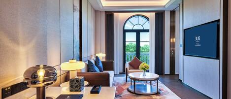 A modern hotel room with a large TV, a sofa, a coffee table, and a view of the outdoors through a large window.