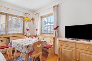 Una habitación con una mesa de madera, sillas, un televisor y ventanas con cortinas.