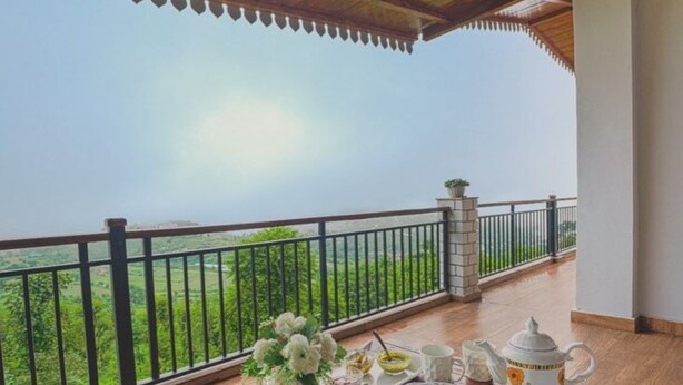 Un balcón con vistas a un paisaje verde, una mesa con juego de té y un arreglo floral.