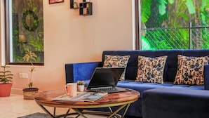 Una sala de estar moderna con un sofá azul, una mesa de centro y una computadora portátil. Hay plantas en macetas y obras de arte en las paredes.