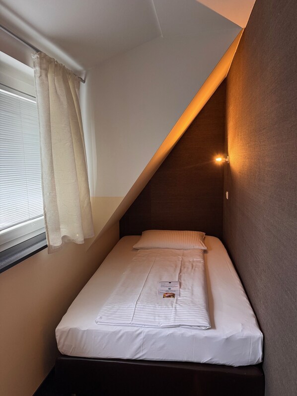 Una habitación pequeña con cama individual, techo inclinado, ventana con cortinas y una lámpara fijada a la pared.