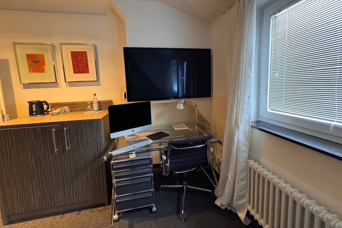 Habitación de hotel compacta con kitchenette, un televisor de pantalla plana en la pared, un escritorio con lámpara y una ventana con persianas.