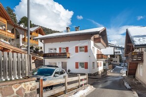 A snow-covered street with wooden houses, a parked car, and a clear blue sky. - Apartment 'Goller Saslonch' with Mountain View, Balcony and Wi-Fi (ORTISEI)
