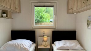 A small, cozy room with two single beds, a window with a view of greenery, and wooden cabinets. - Furnished Chalet in Sint Maartenszee (Sint Maartensvlotbrug)