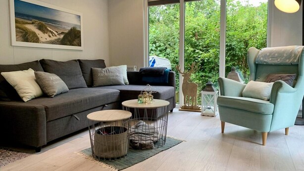 A modern living room with a grey sofa, a teal armchair, and a round coffee table. There is a large window with a view of greenery outside. - Furnished Chalet in Sint Maartenszee (Sint Maartensvlotbrug)