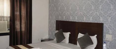 Un lit bien fait, recouvert d’une couvre-lit à rayures, avec une tête de lit sombre et deux oreillers. On y trouve une petite table de chevet avec une lampe et une bouteille d’eau. La pièce est décorée d’un papier peint à motifs et un tableau encadré orne le mur.