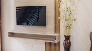 A flat-screen TV mounted on a wall with a wooden panel design, a shelf below it, and a bamboo plant in a vase to the right. - Elyseah Condotel Angeles Pampanga (Angeles City)